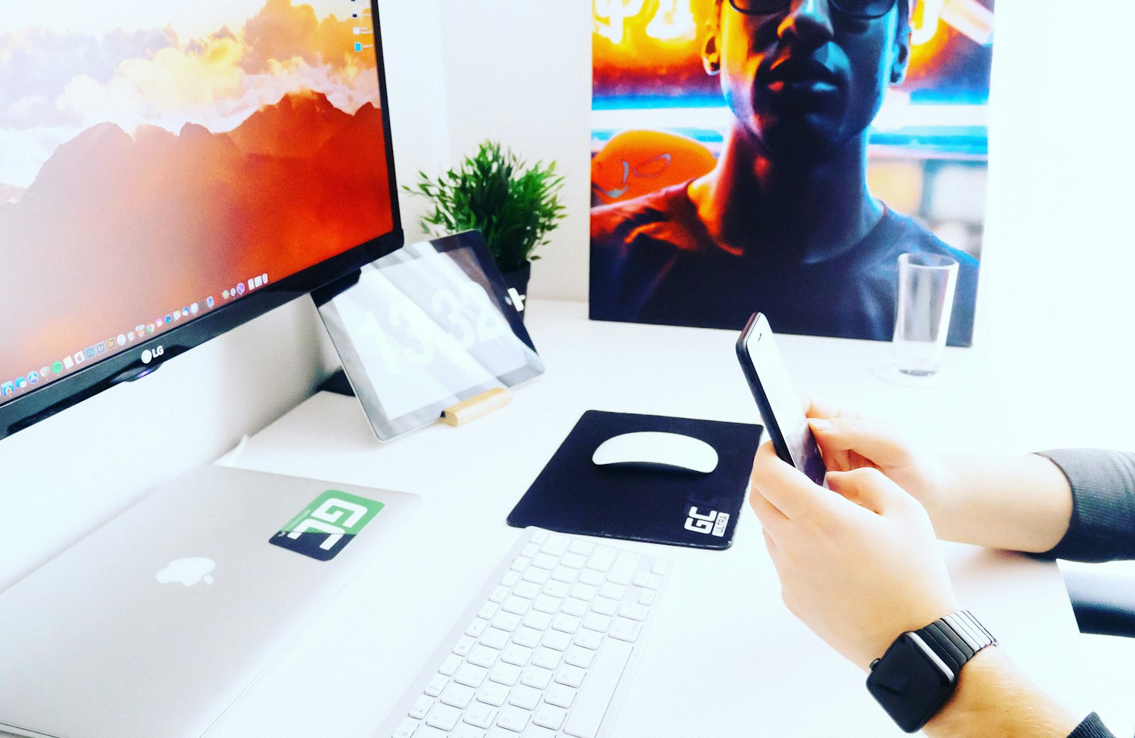 A phone in hand next to a desktop monitor and laptop on a white desk